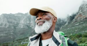 View, face or senior black man on mountain hiking in outdoor adventure or journey in nature for tra